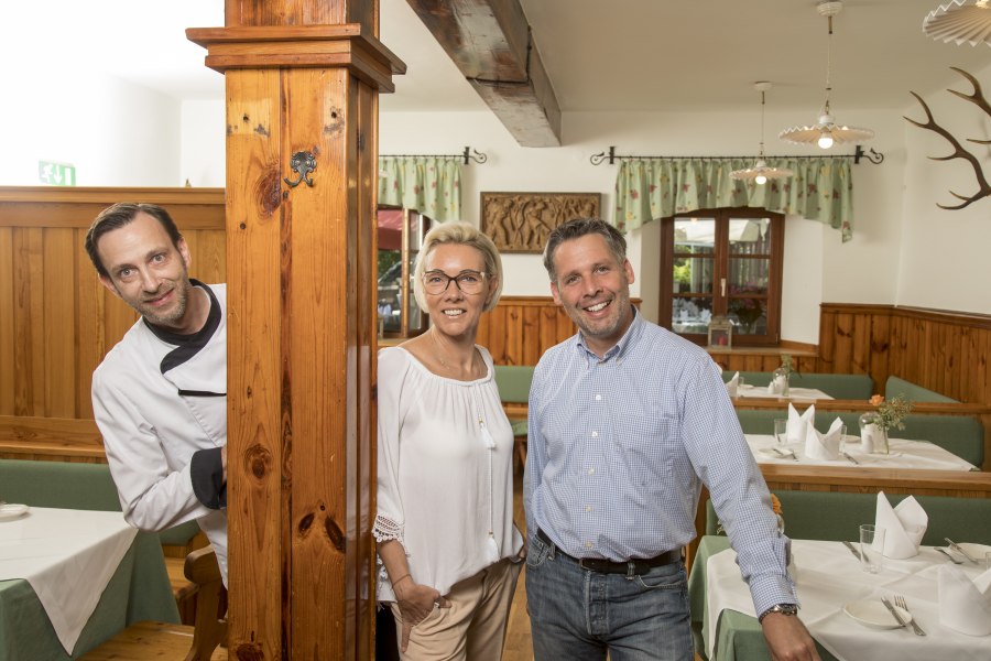 Drei Personen in einem rustikalen Restaurant, eine Person in Kochkleidung, zwei in Freizeitkleidung, lächeln in die Kamera.
