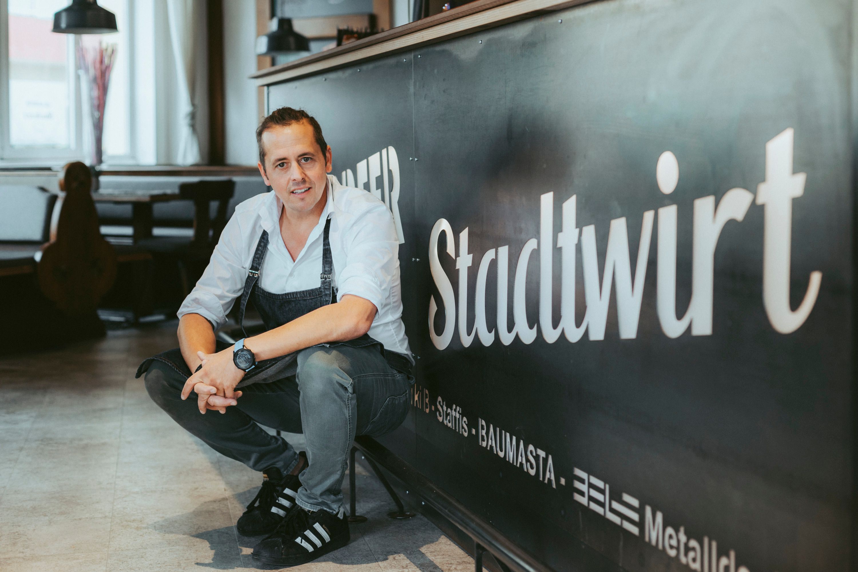 Ein Mann in Arbeitskleidung hockt vor einem Schild mit der Aufschrift 'Stadtwirt'.