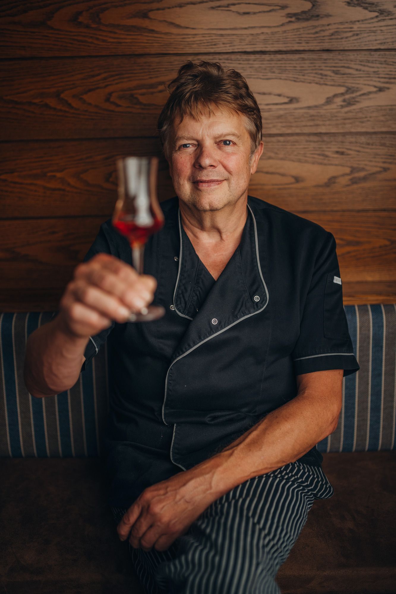 Ein Mann in Kochkleidung hält ein Glas mit rotem Getränk vor einer Holzwand.