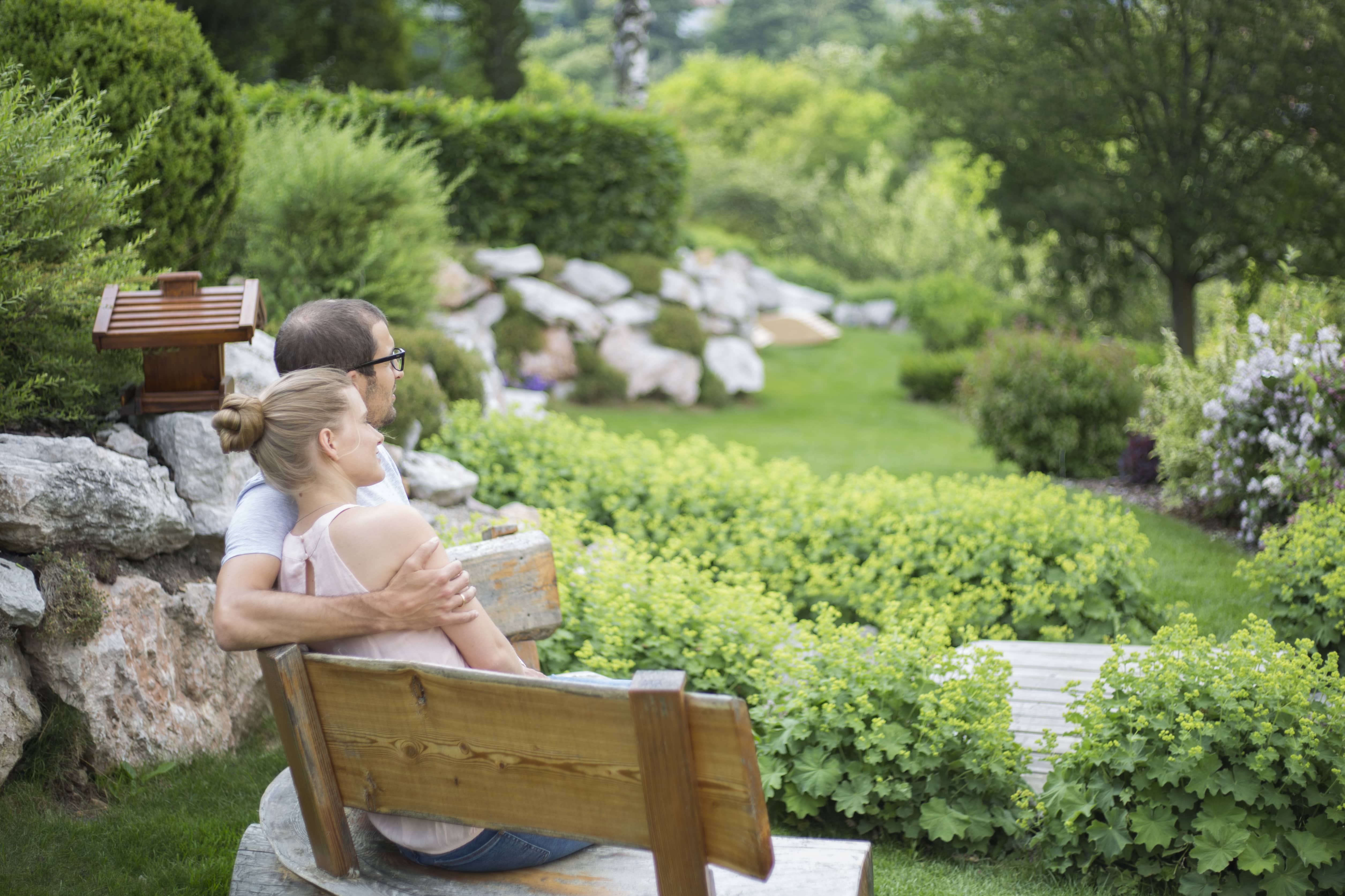 Ein Paar sitzt auf einer Holzbank in einem grünen Garten mit Büschen und Bäumen.