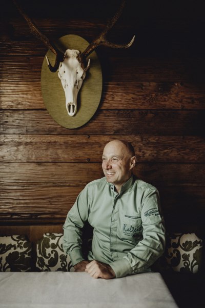 Ein Mann in einem gr&uuml;nen Hemd sitzt an einem Tisch vor einer Holzwand mit einem Hirschgeweih.