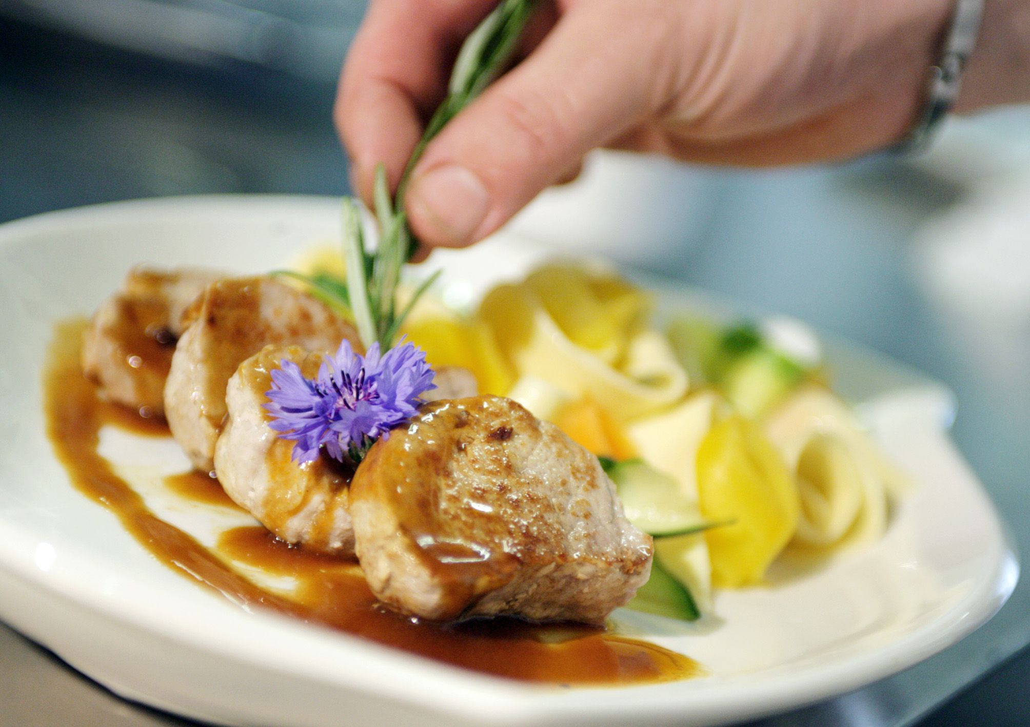 Ein Teller mit Fleisch, Soße, Nudeln und Gemüse, dekoriert mit einer Blume und Rosmarin.