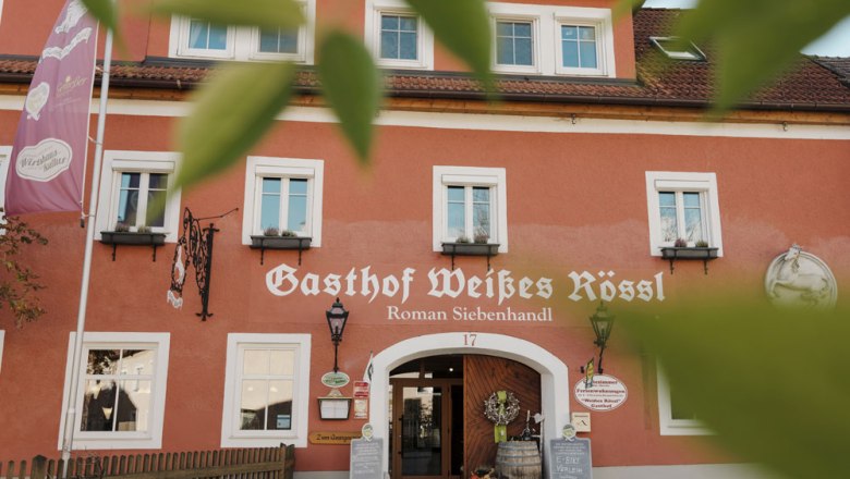 Fassade des Gasthofs Wei&szlig;es R&ouml;ssl in M&uuml;hldorf mit roter Wand und wei&szlig;en Fenstern.