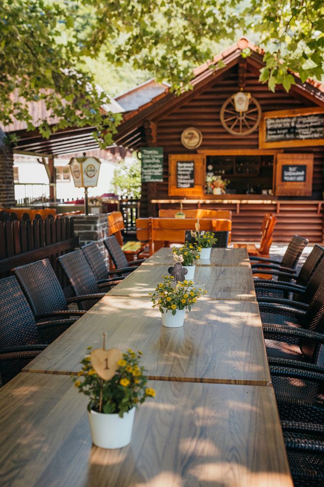 Ein langer Holztisch im Freien mit Blumen in Töpfen, umgeben von Stühlen. Im Hintergrund eine Holzhütte mit Schildern.