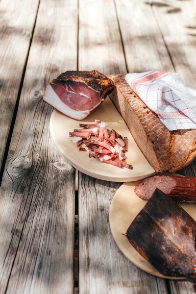 Brot, Speck und Wurst auf Holztisch im Freien angerichtet.
