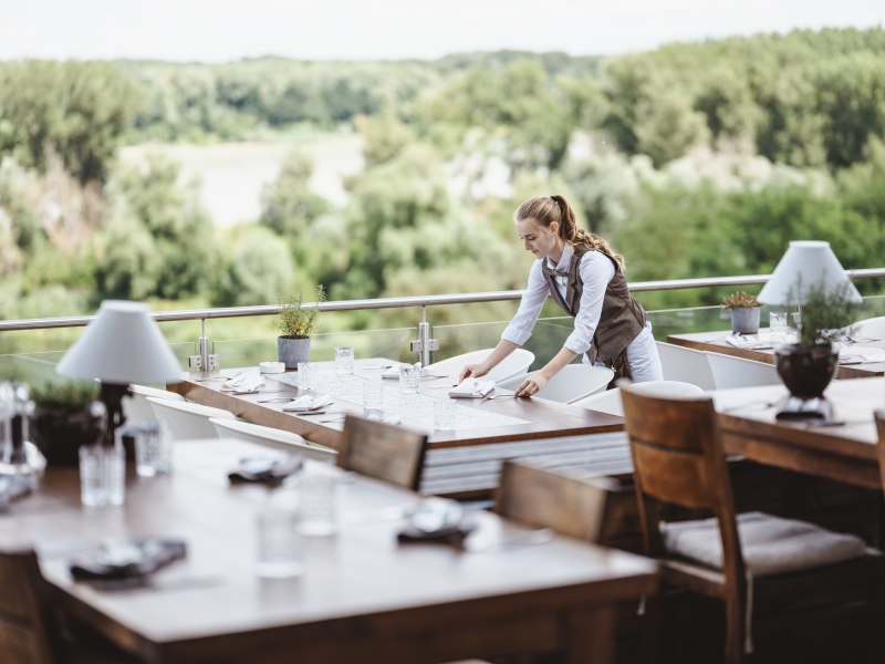 Eine Kellnerin deckt Tische auf einer Terrasse mit Blick auf gr&uuml;ne W&auml;lder.