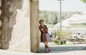 Frau lehnt lächelnd an einer Mauer, im Hintergrund Terrasse mit Tischen, Stühlen und Sonnenschirmen.
