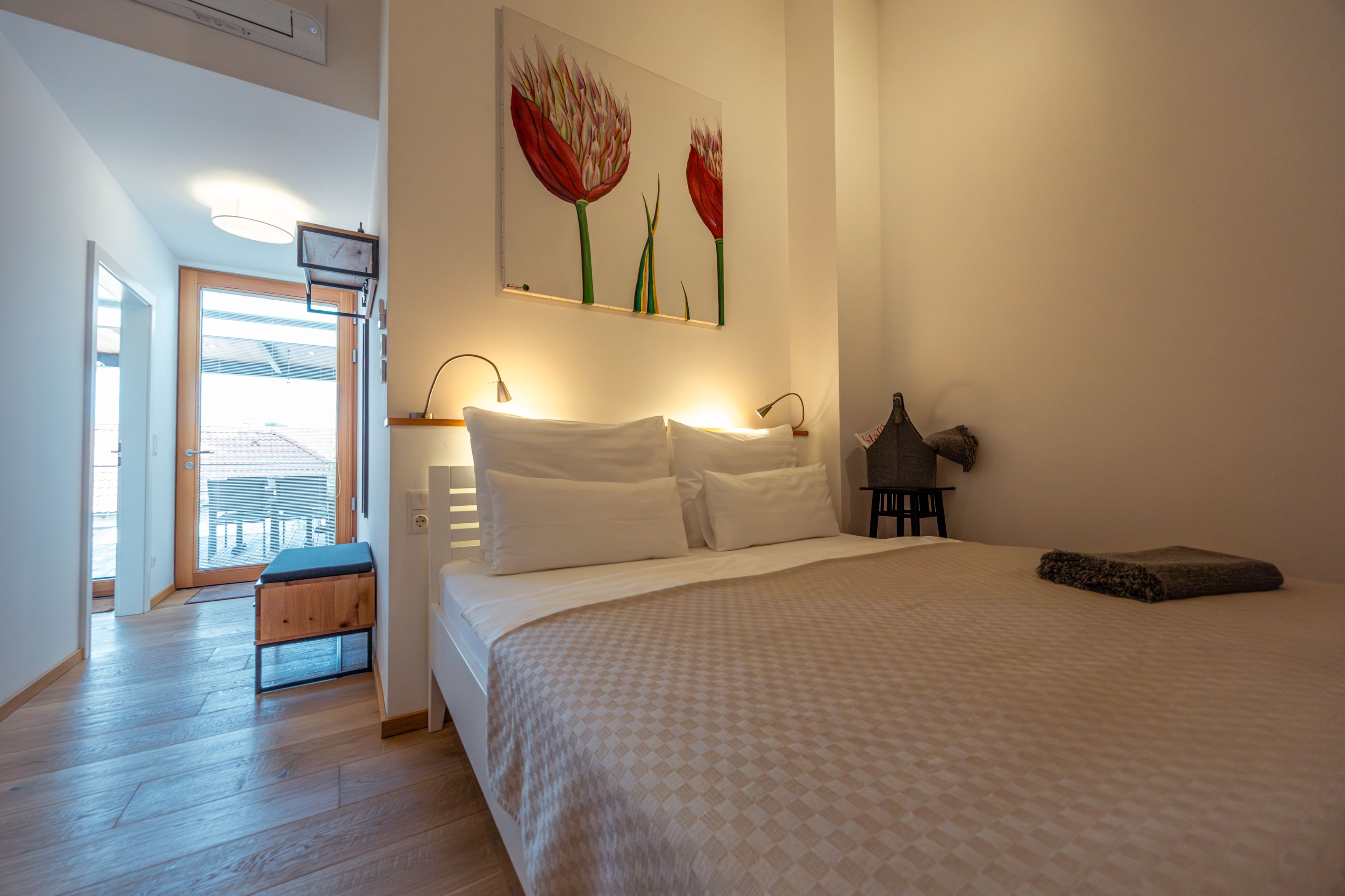 Modern bedroom with double bed, light wooden floor and artwork on the wall. Door leads to a balcony with views of rooftops.