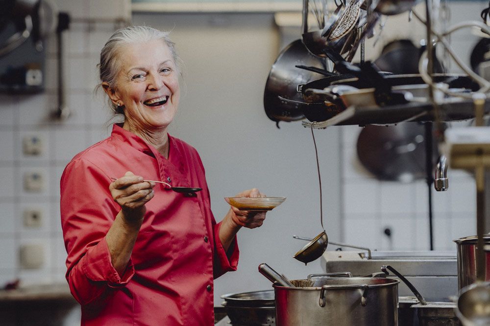 A smiling woman in a red chef's jacket stands in a kitchen with a spoon and a bowl in her hand.