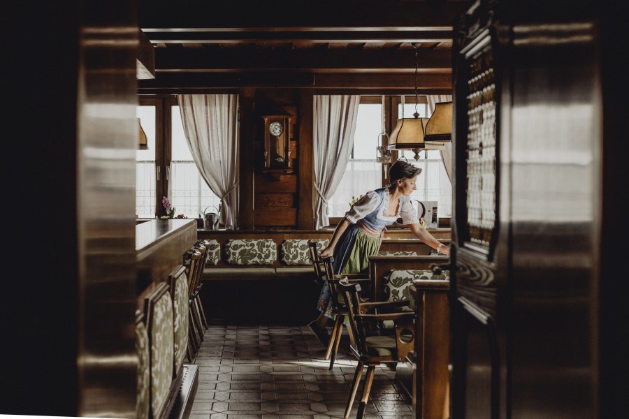 Eine Frau in traditioneller Kleidung richtet einen Tisch in einem gem&uuml;tlichen, holzget&auml;felten Gastraum her.