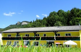 Gelbes Geb&auml;ude mit Blumenbalkon vor bewaldetem H&uuml;gel und blauem Himmel.