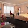Zwei Frauen in einem Hotelzimmer mit Doppelbett, eine sitzt, die andere steht mit erhobenem Glas.