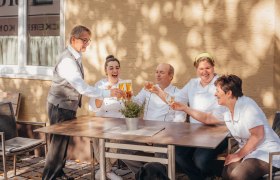 Ein Team von f&uuml;nf Personen sitzt drau&szlig;en an einem Tisch und st&ouml;&szlig;t mit Getr&auml;nken an. Ein schwarzer Hund sitzt davor.