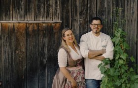 Ein Paar steht vor einer Holzwand, sie trägt ein Dirndl, er eine Kochjacke.