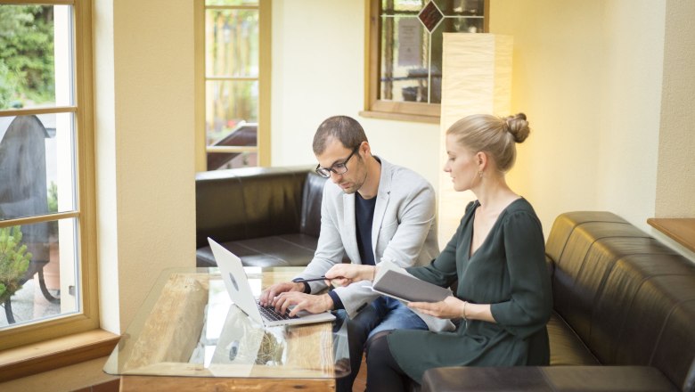 Zwei Personen arbeiten an einem Laptop in einem gem&uuml;tlichen Raum mit gro&szlig;en Fenstern.