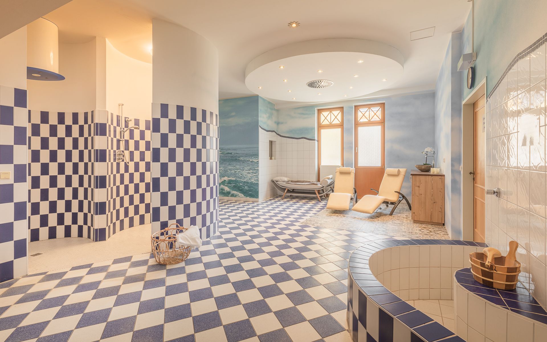 Innenansicht eines Wellnessbereichs mit blauen und weißen Fliesen, Liegestühlen und einem Holzeimer.