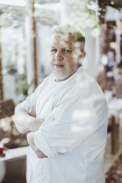 Ein Mann in Kochkleidung steht mit verschr&auml;nkten Armen hinter einer Glasscheibe.
