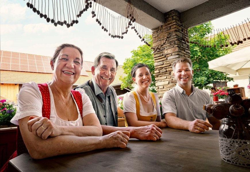 Vier Personen sitzen lächelnd an einem Tisch im Freien, umgeben von Blumen und einem rustikalen Ambiente.