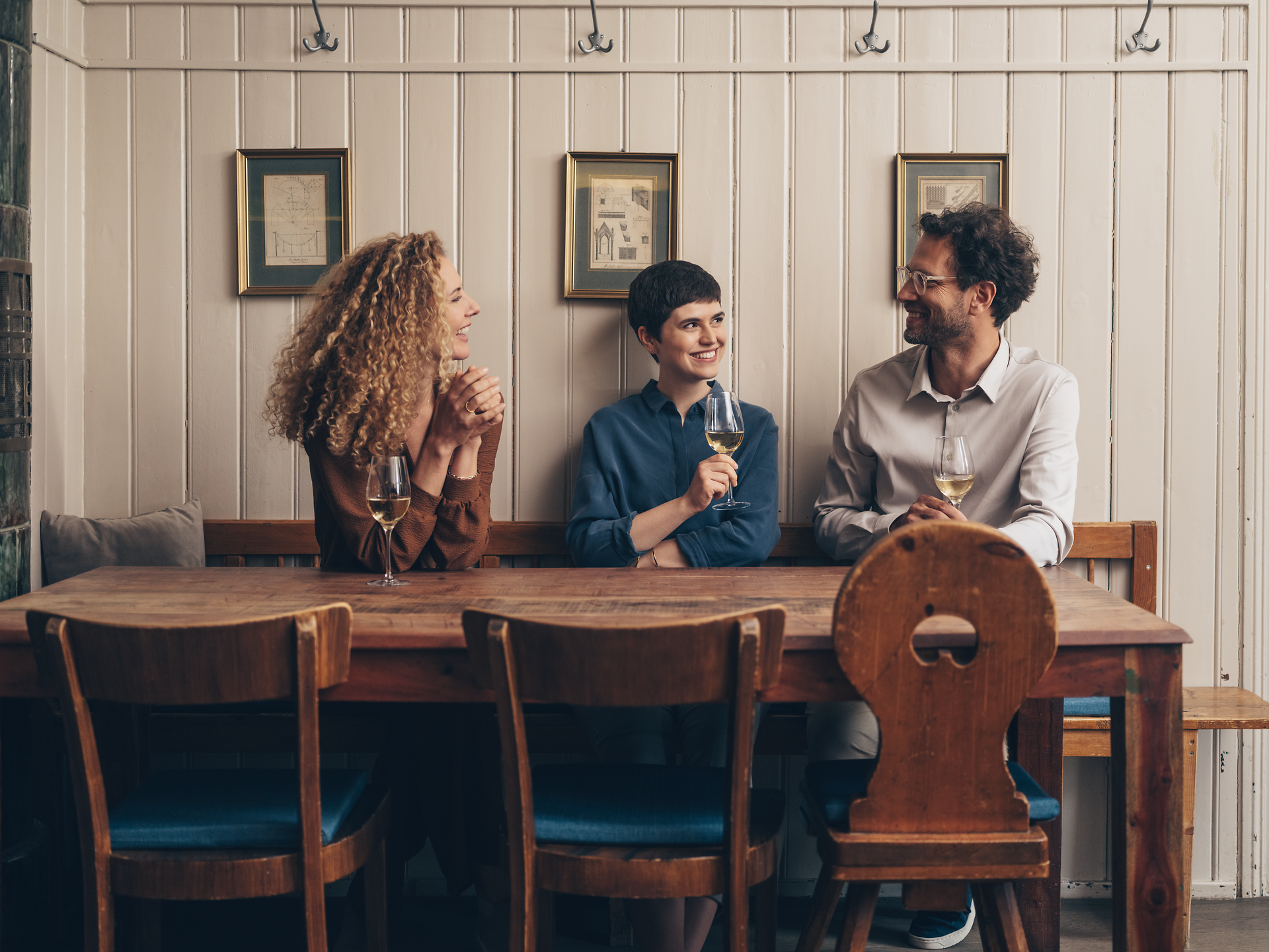 In einem einladenden Wirtshaus genießen drei Freunde angeregte Gespräche bei einem Glas Wein. Die warme Atmosphäre und die rustikale Einrichtung schaffen einen perfekten Rahmen für gesellige Stunden und kulinarische Entdeckungen in der Region.