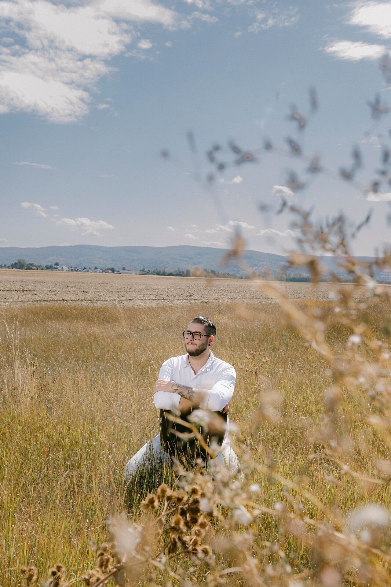 Eine Person mit Brille und weißem Hemd sitzt mit verschränkten Armen in einer Wiese. Im Hintergrund sind Felder, Hügel und ein blauer Himmel zu sehen.
