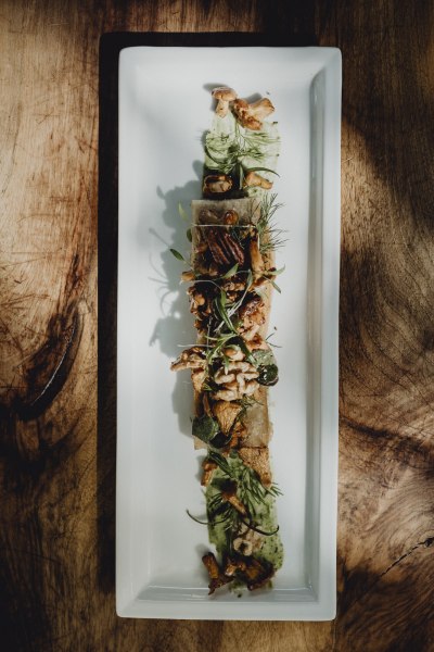 Ein rechteckiger Teller mit sautierten Eierschwammerln, Pecann&uuml;ssen, Waldkr&auml;utern und Wurzelbrot, kunstvoll angerichtet auf einem Holztisch.