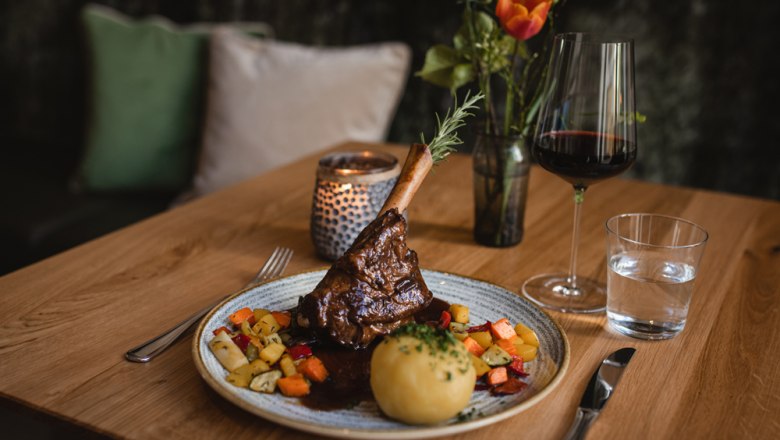 Ein Teller mit Lammstelze, Kn&ouml;del und Gem&uuml;se, daneben ein Glas Rotwein und Wasser auf einem Holztisch.
