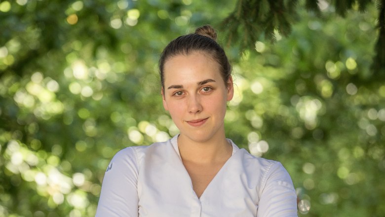 Eine Frau in wei&szlig;em Hemd steht mit verschr&auml;nkten Armen vor einem unscharfen, gr&uuml;nen Hintergrund.