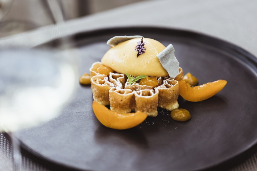 Eine kunstvoll angerichtete Waffel mit Marillensorbet und dekorativen Früchten auf einem schwarzen Teller.