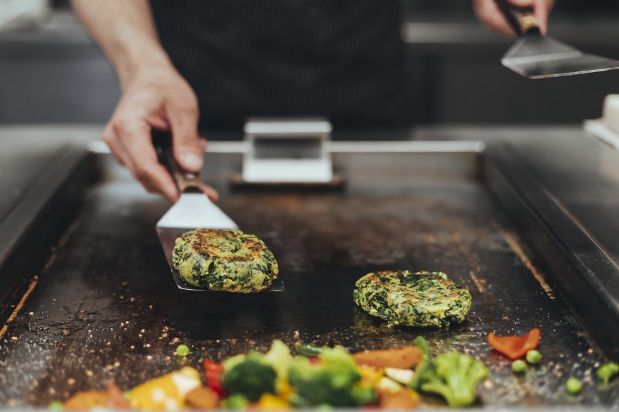 Ein Koch wendet Gem&uuml;sebratlinge auf einer Grillplatte, umgeben von buntem Gem&uuml;se.