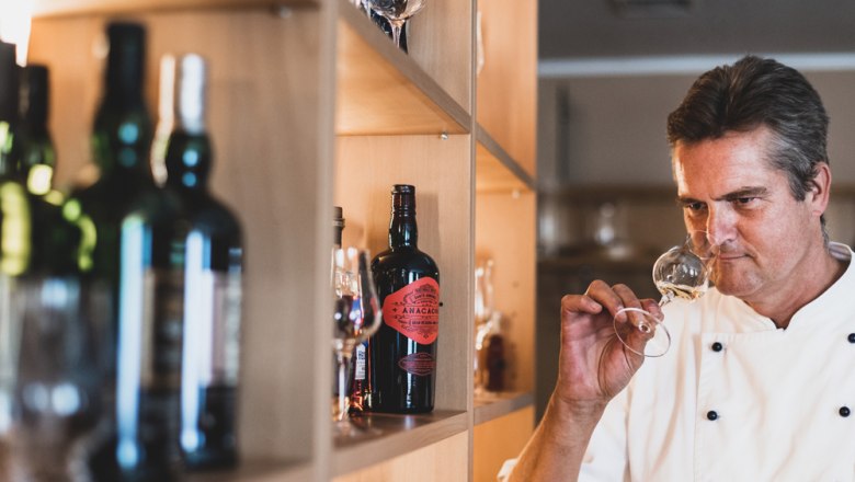 Ein Mann in Kochjacke riecht an einem Weinglas vor einem Regal mit Flaschen.