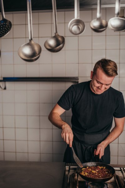 Ein Koch in schwarzer Kleidung br&auml;t in einer Pfanne in einer K&uuml;che mit h&auml;ngenden Kellen.