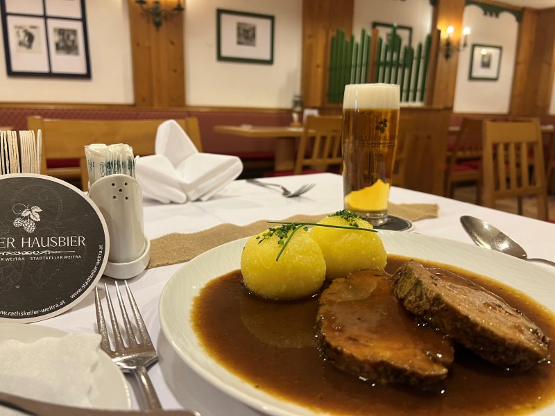 Ein Teller mit Schweinsbraten und zwei Erdäpfelknödeln in einem Restaurant, daneben ein Glas Bier.