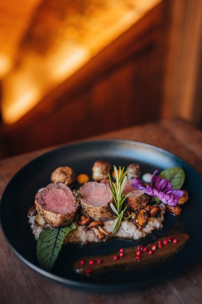Ein Teller mit rosa gebratenem Hirschsteak auf Risotto, garniert mit Rosmarin, Pilzen und einer Blüte.
