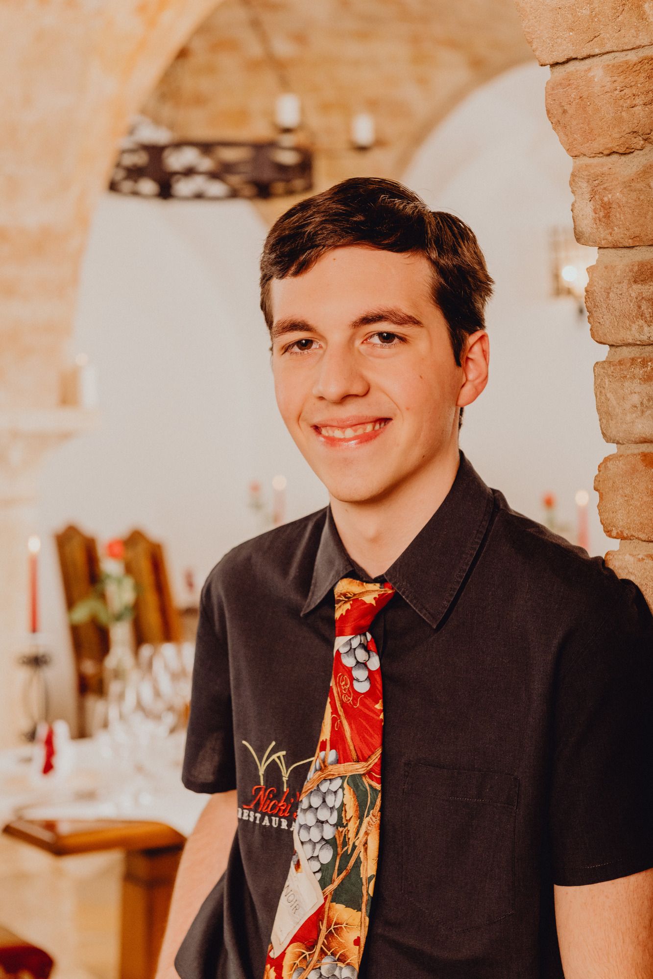 Ein junger Mann in einem Restaurant mit einer bunten Krawatte, lehnt an einer Backsteinmauer.