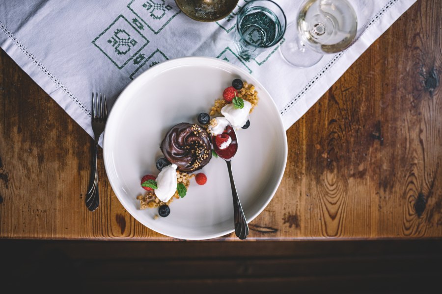 Ein Dessertteller mit einem Nougat-T&ouml;rtchen, Hollereis, Beeren und einem L&ouml;ffel auf einem Holztisch.