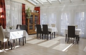 Ein heller, eleganter Speisesaal mit gedeckten Tischen und St&uuml;hlen, umgeben von gro&szlig;en Fenstern mit wei&szlig;en Vorh&auml;ngen.