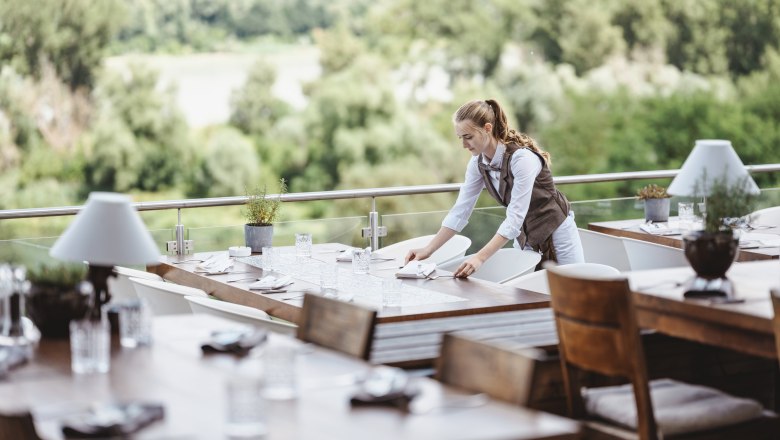 Eine Kellnerin deckt Tische auf einer Terrasse mit Blick auf gr&uuml;ne W&auml;lder.