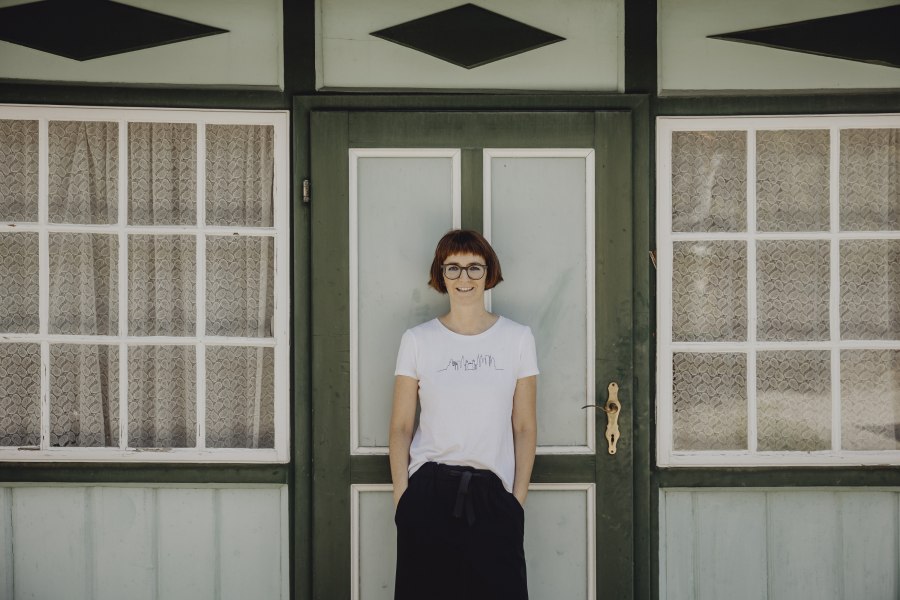 Eine Frau steht vor einer gr&uuml;nen T&uuml;r mit Fenstern und Spitzenvorh&auml;ngen.