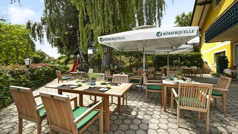 Ein sonniger Gastgarten mit Holzmöbeln und Sonnenschirmen, umgeben von grüner Vegetation und einem gelben Gebäude.