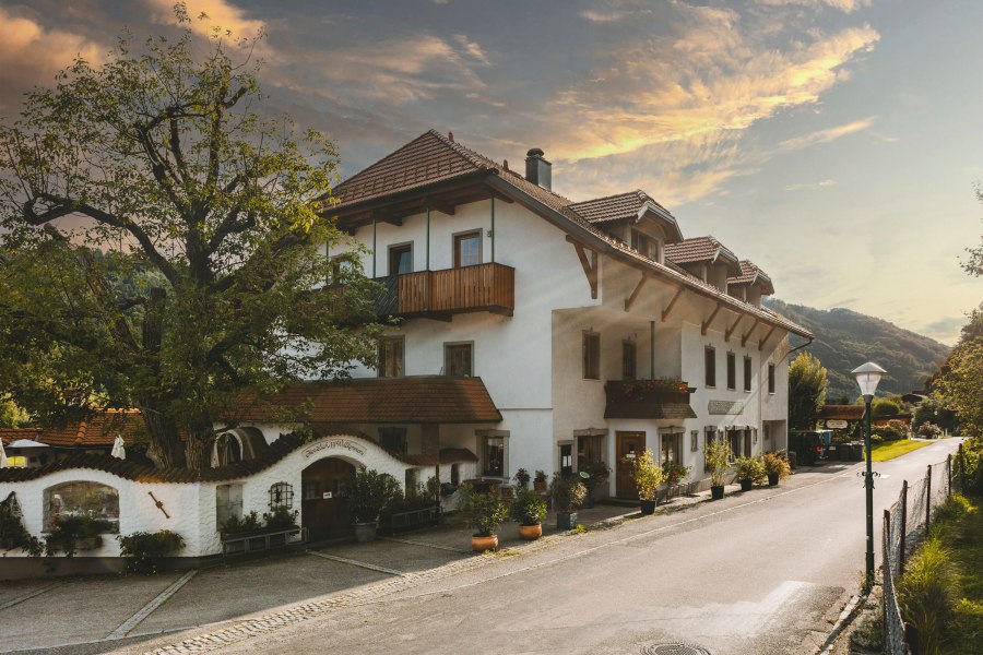 Ein traditionelles Wirtshaus mit weißen Wänden und roten Ziegeldächern, umgeben von Bäumen und einer ruhigen Straße.
