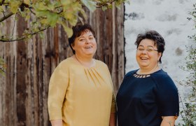 Zwei lachende Frauen stehen vor einer Holzwand im Freien.