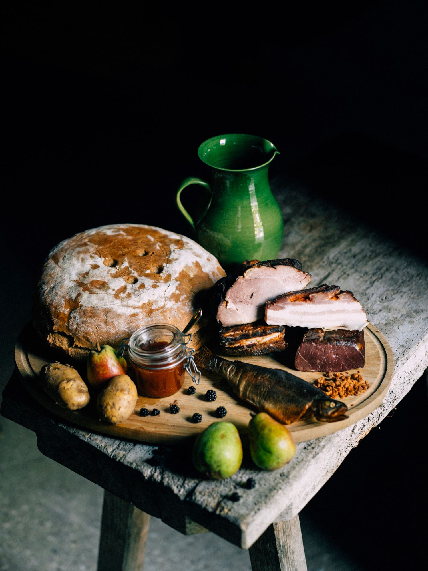 Ein rustikales Arrangement von Brot, Fleisch, Fisch, Birnen, Kartoffeln und einem grünen Krug auf einem Holztisch.