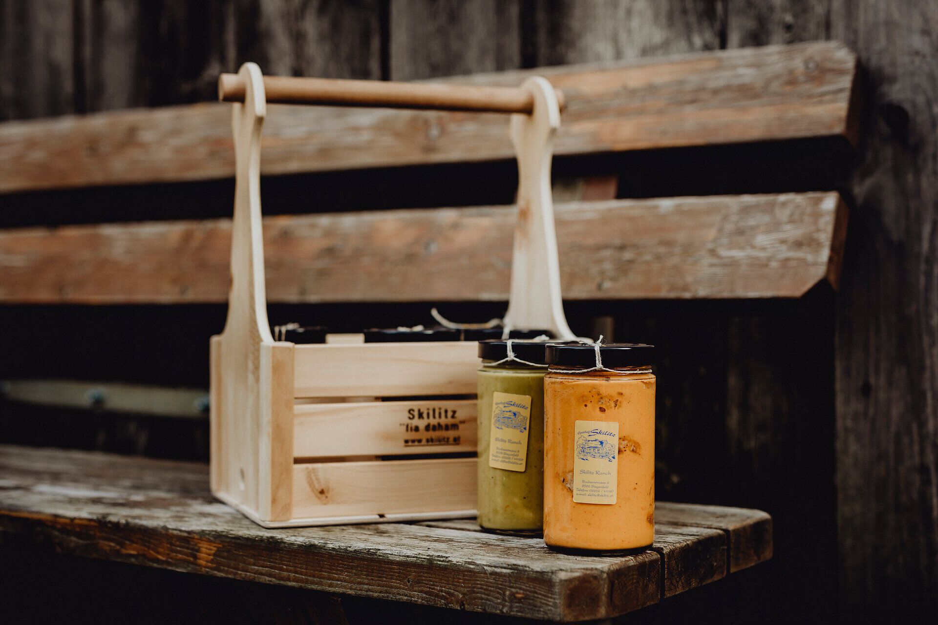 Inmitten der malerischen Landschaft des Wienerwaldes laden die bunten Gläser mit köstlichen Speisen zum Genießen ein. Die rustikale Holzbank und die charmante Männerhandtasche schaffen eine einladende Atmosphäre, die zum Verweilen einlädt. Hier wird die Wirtshauskultur Niederösterreichs auf besondere Weise zelebriert.