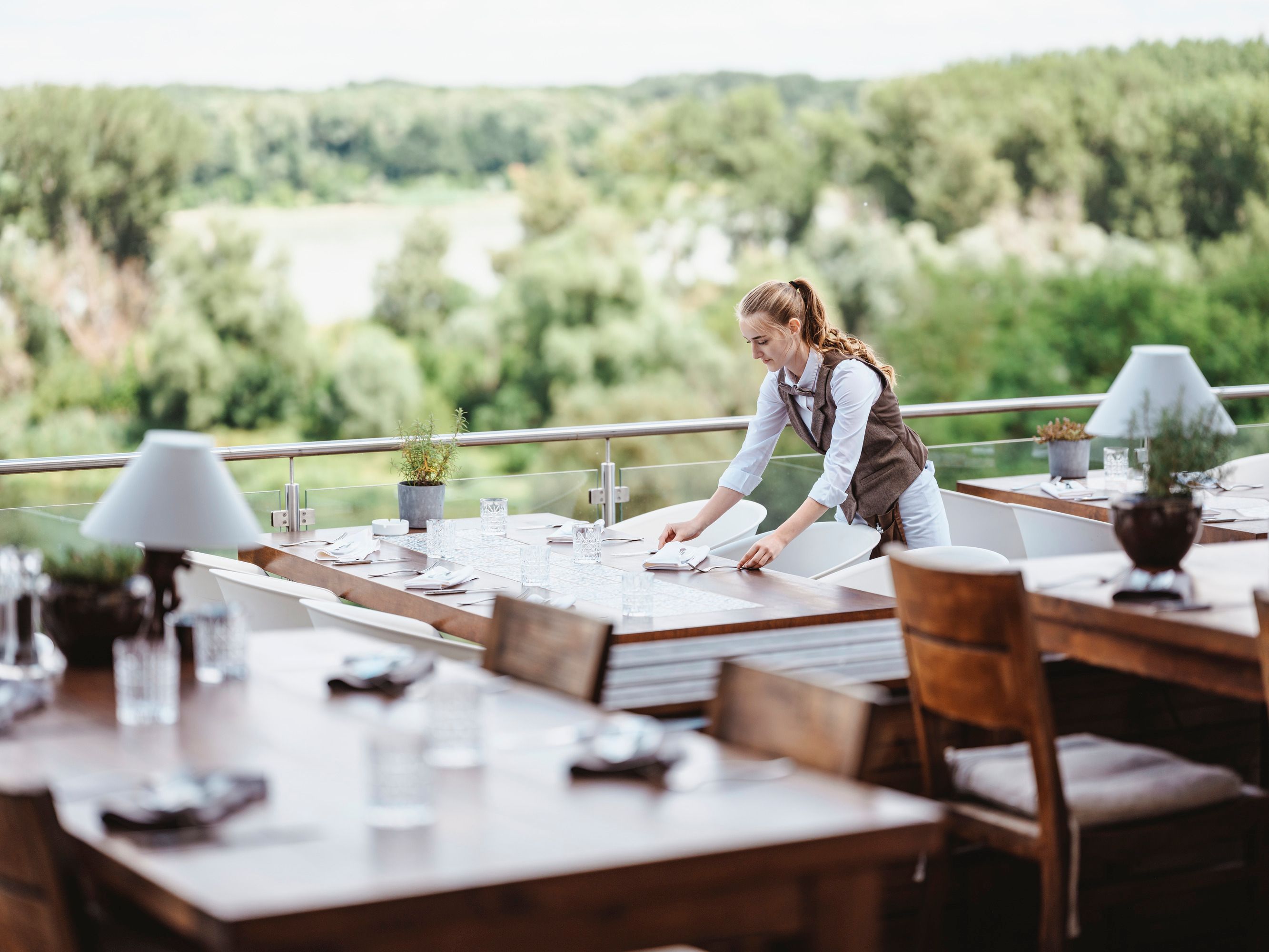 Eine Kellnerin deckt Tische auf einer Terrasse mit Blick auf grüne Wälder.
