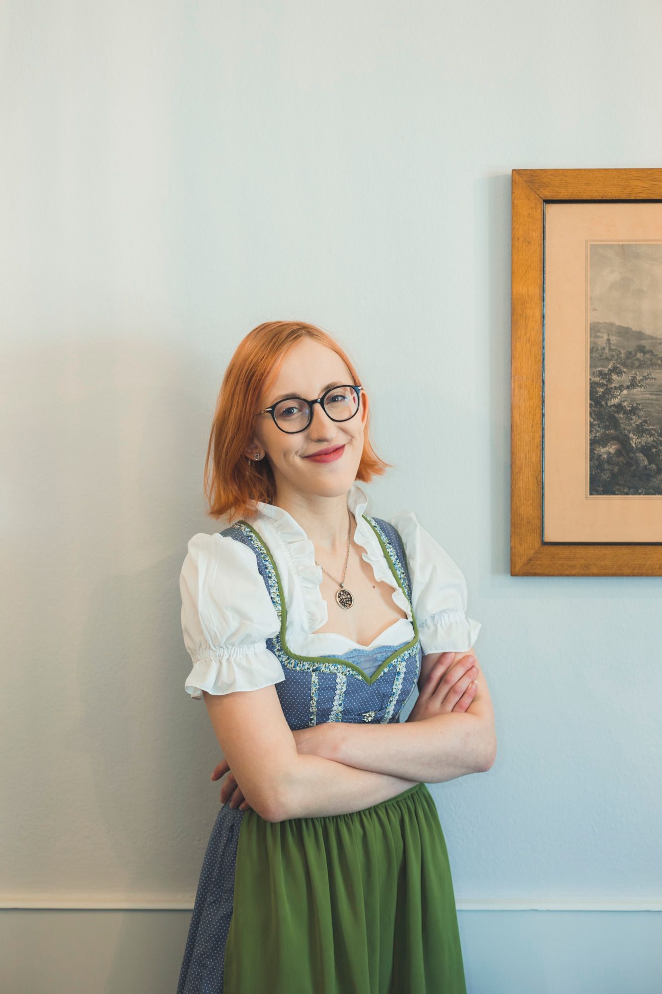 Eine Frau mit Brille und rotem Haar trägt ein traditionelles Dirndl und steht lächelnd vor einer weißen Wand neben einem gerahmten Bild.