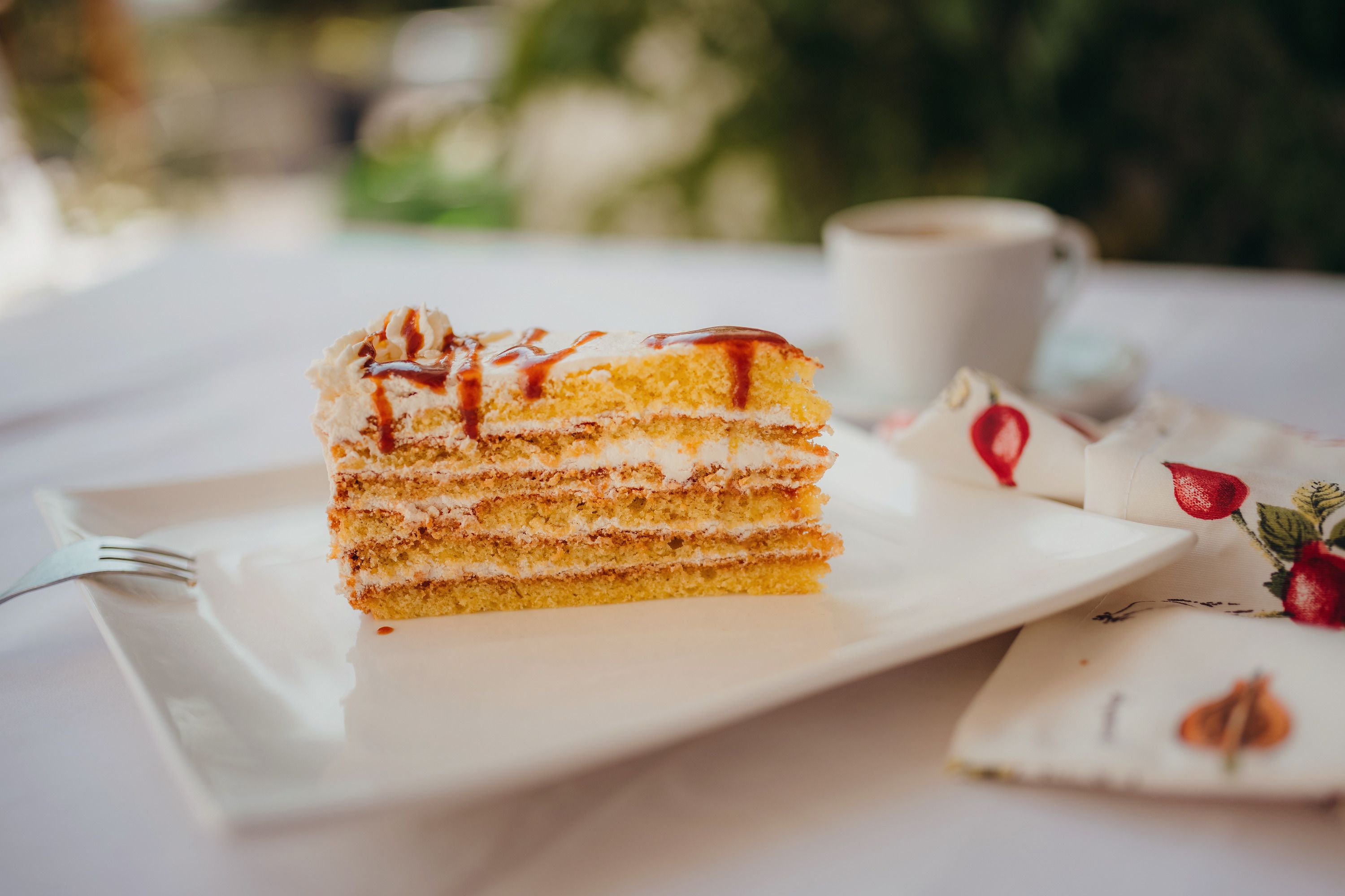 Ein Stück Schichttorte mit Sahne und Karamell auf einem weißen Teller, daneben eine Tasse Kaffee und eine Serviette mit Fruchtmuster.