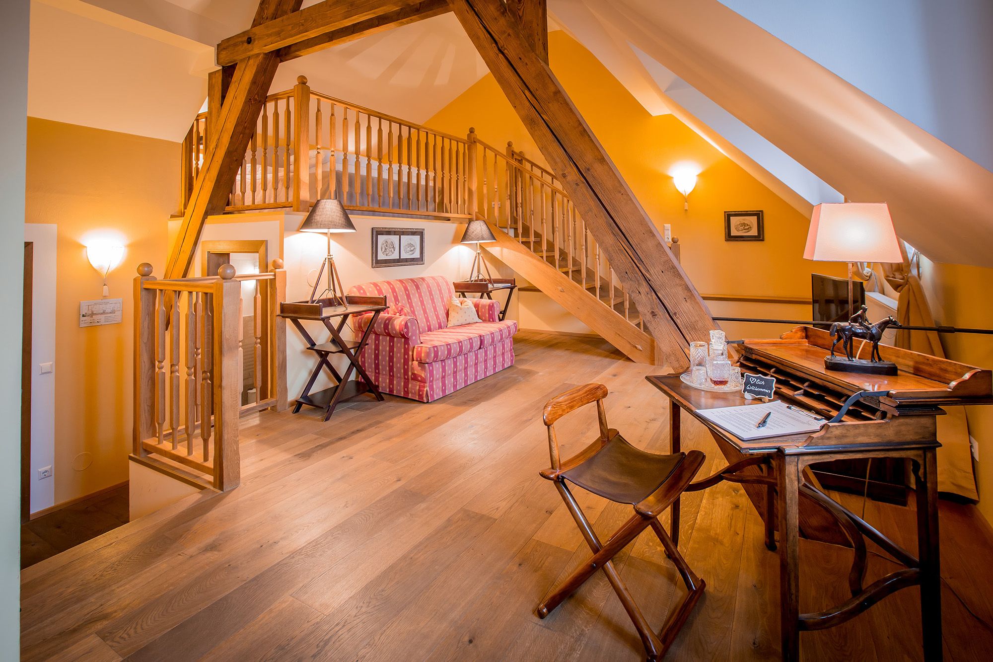 Gemütliche Dachgeschoss-Suite mit Holzbalken, Sofa, Schreibtisch und Treppe zu einem Schlafbereich.
