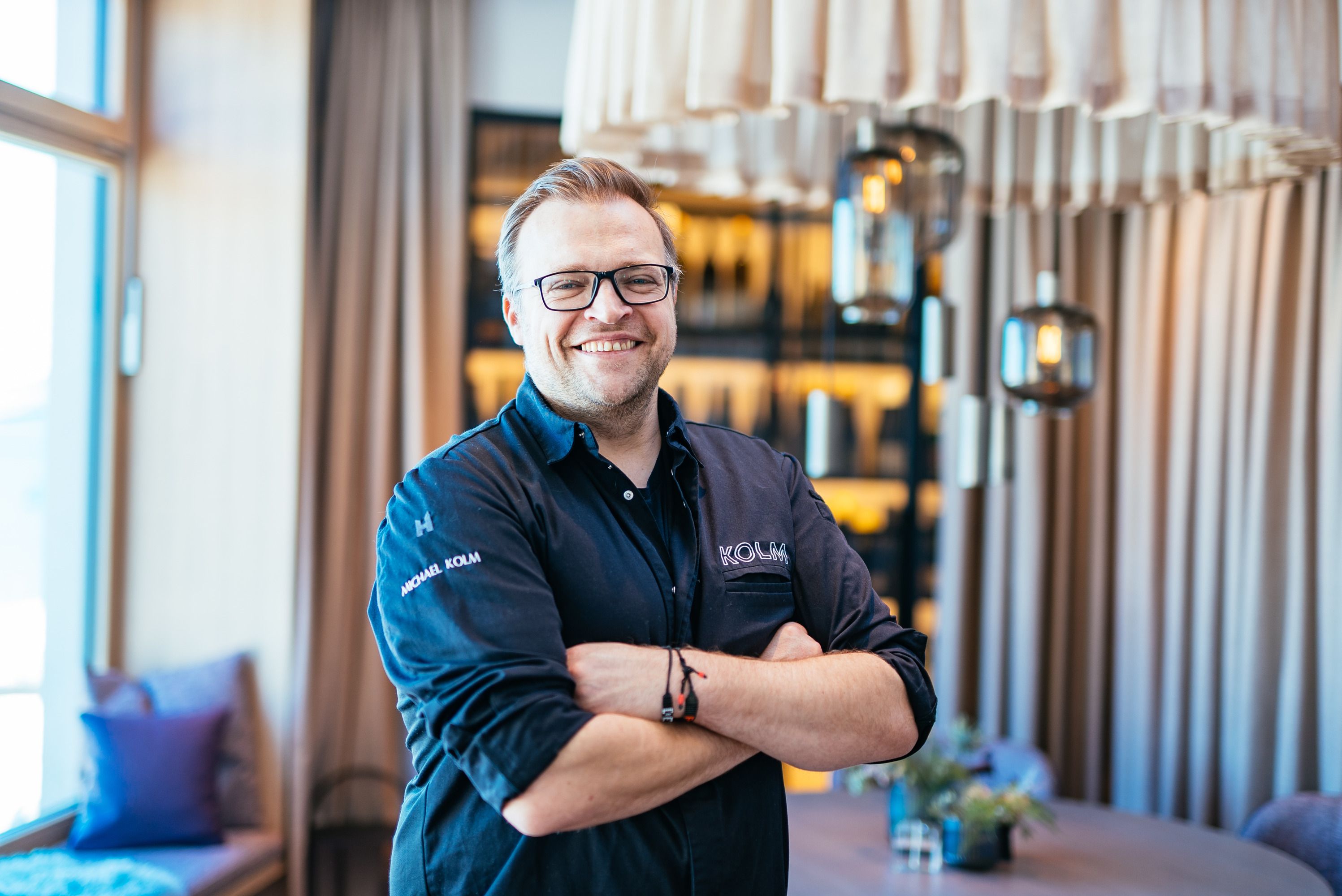 Ein lächelnder Mann in Kochkleidung steht mit verschränkten Armen in einem stilvollen Restaurant.