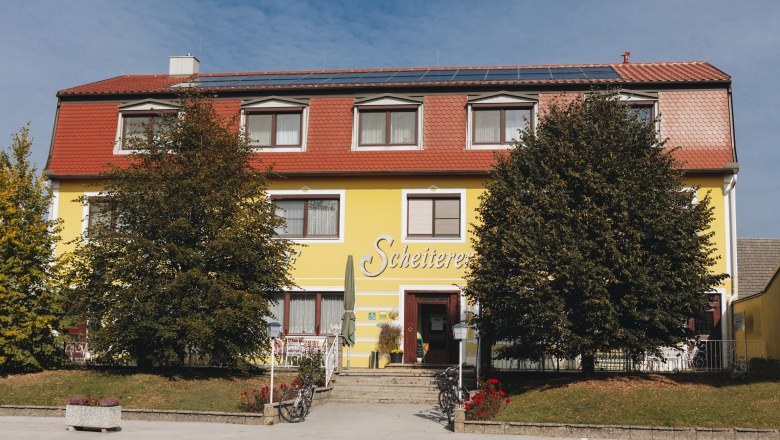 Gelbes Gebäude mit rotem Dach und der Aufschrift 'Scheiterer', umgeben von Bäumen und Fahrrädern.