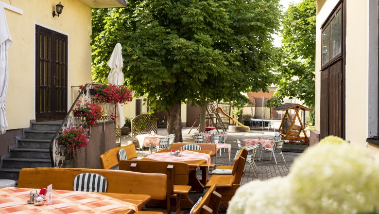 Ein schattiger Gastgarten mit Holztischen und Stühlen, umgeben von Bäumen und Blumen, ideal für heiße Sommertage.